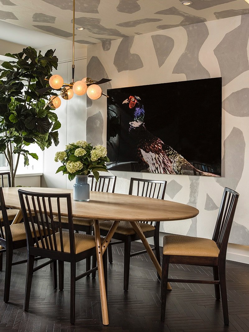 A contemporary dining room with abstract gray and white patterned walls and ceiling, an oval light wood table, dark ladder-back chairs, and a large, colorful pheasant painting.