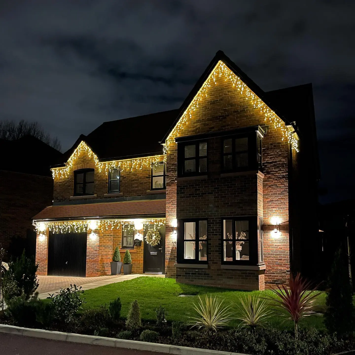 A two-story brick house with dark trim is brightly decorated with thick, warm white icicle lights strung along the rooflines of the gables, giving a heavy, glowing border.