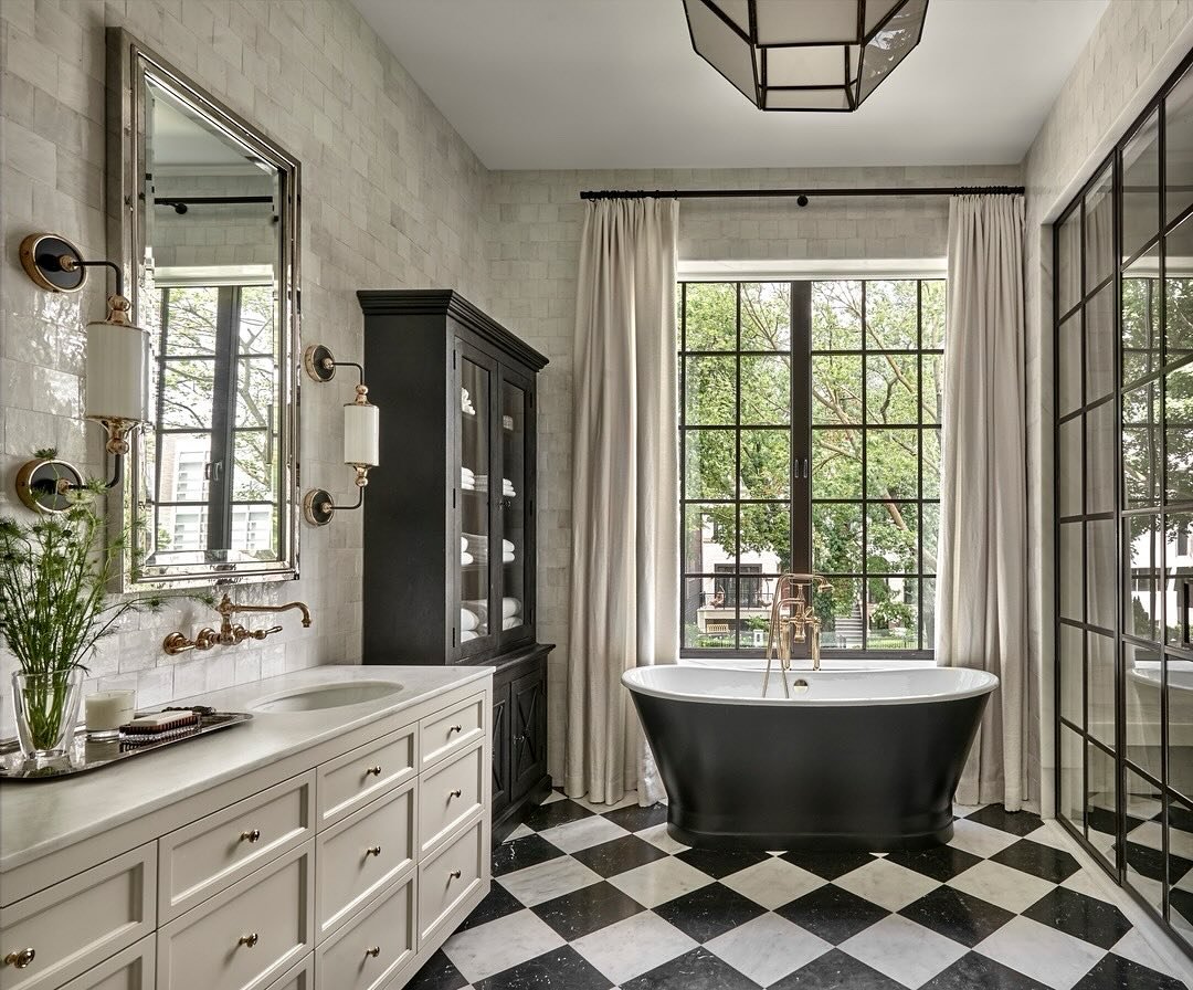 A dramatic, luxurious bathroom with a black freestanding tub, a white double vanity, and a floor of high-contrast black and white marble tiles in a diamond pattern.