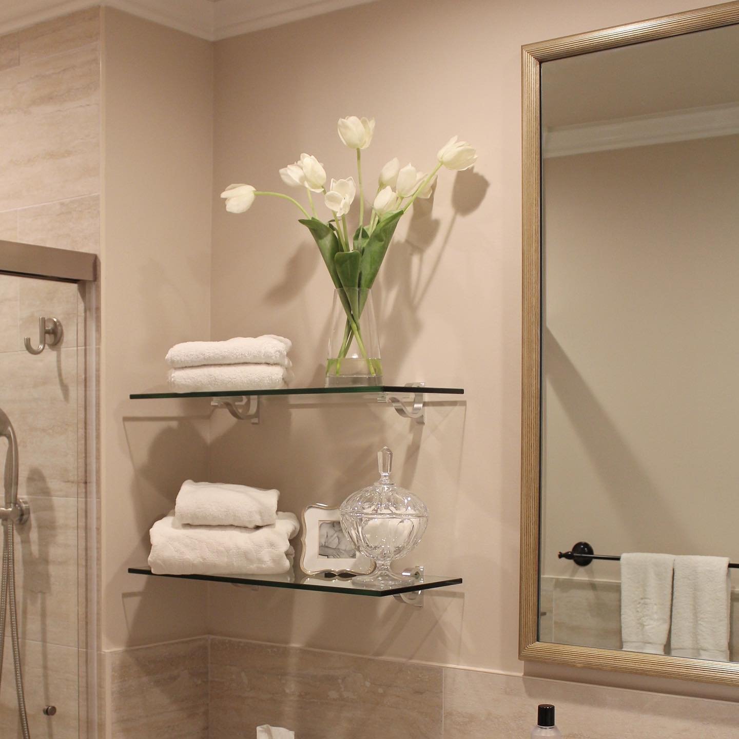 Two clear glass shelves supported by silver brackets, holding white folded towels, a clear vase with white tulips, and a crystal jar next to a framed mirror.