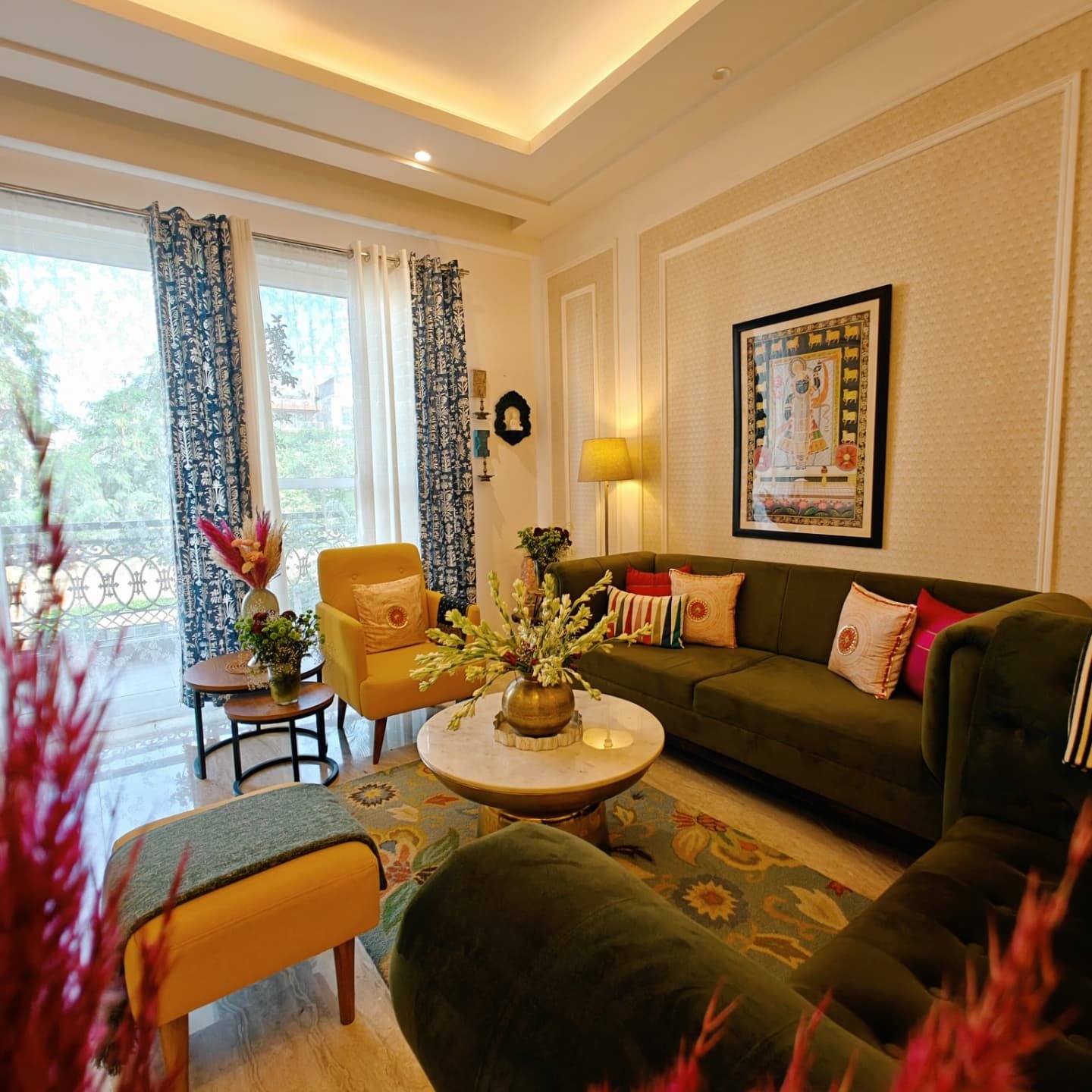 Vibrant living room with a deep olive green velvet sofa, mustard yellow accent chairs, and a framed traditional Indian artwork.