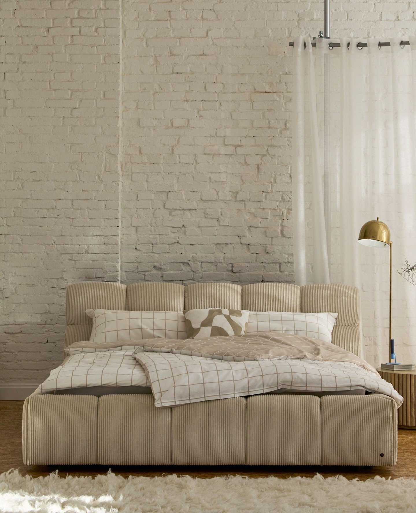 An industrial-style beige bedroom featuring a large, vertically ribbed corduroy upholstered bed and headboard, checkered bedding, a brass floor lamp, and a white painted brick wall.