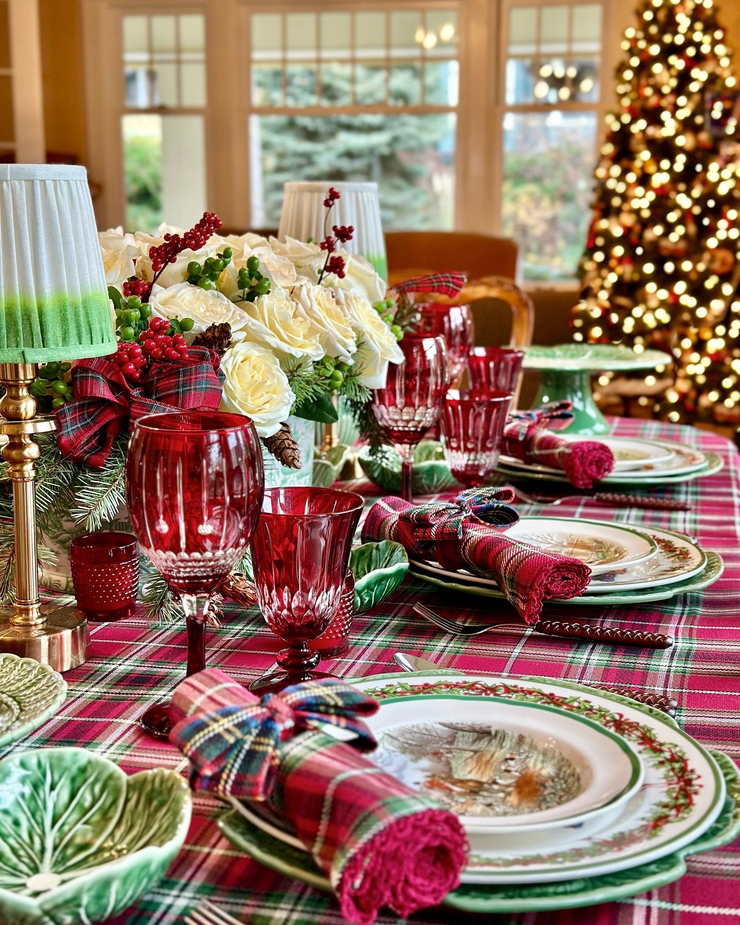 A festive dining table covered in a red and green plaid tartan tablecloth, set with layered plates including green rimmed china, plaid rolled napkins tied with ribbon, red cut-glass goblets, and a centerpiece of white roses and red berries.