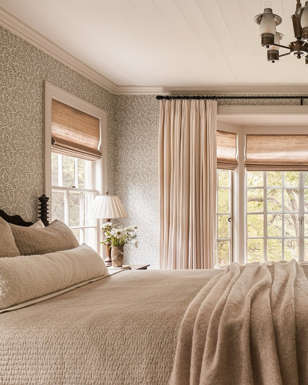 Cream pinch-pleat curtains and natural woven wood shades in a bedroom with patterned walls.