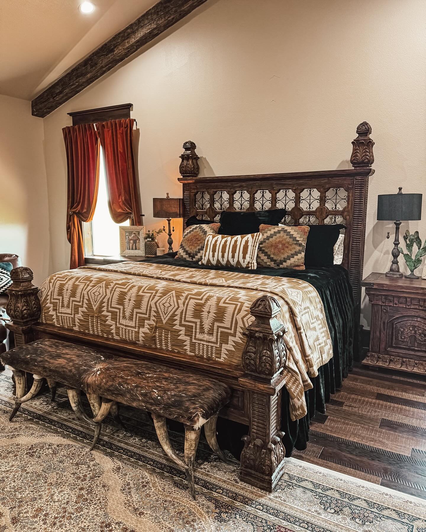 A luxurious Western bedroom with a dark, elaborately carved wood bed, velvet bedding, a beam ceiling, and a cowhide bench with curved horns for legs.