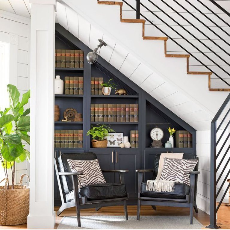 Dark blue under-stairs shelving for books and two black armchairs creating a cozy library corner.