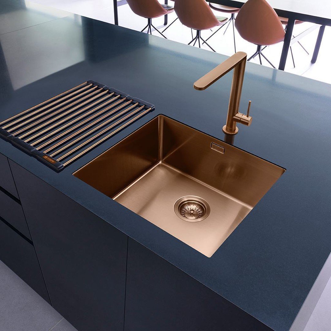 A contemporary single-bowl kitchen sink with a unique PVD copper/rose gold finish, undermounted in a dark matte blue or black countertop, paired with a matching rose gold faucet.