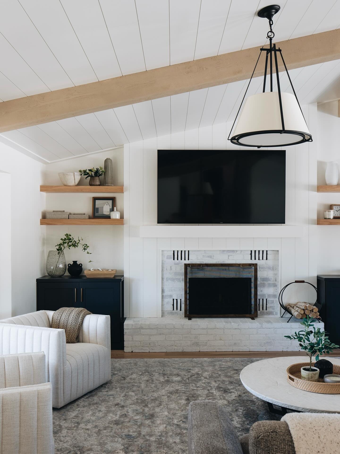 Living room with a white painted brick fireplace, shiplap chimney breast, black accent cabinets, and floating wood shelves.