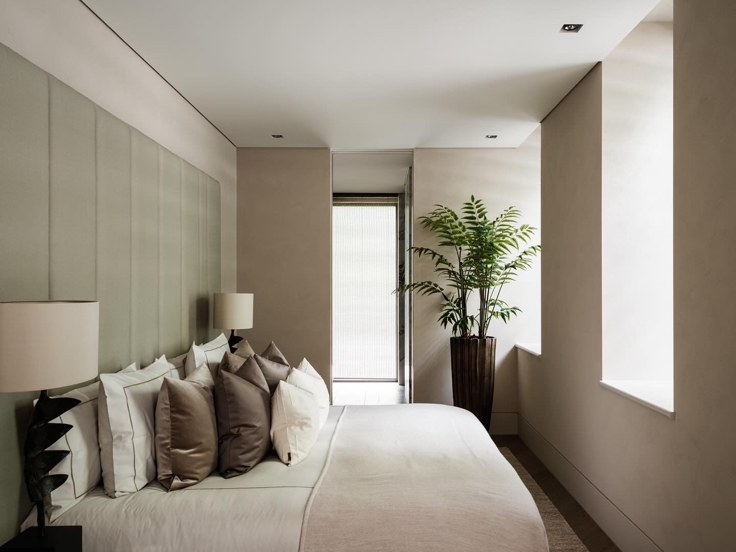 A high-end, contemporary beige bedroom with a pale green padded headboard, layered pillows in white and rich taupe velvet, and a tall potted plant in a dark wood planter.