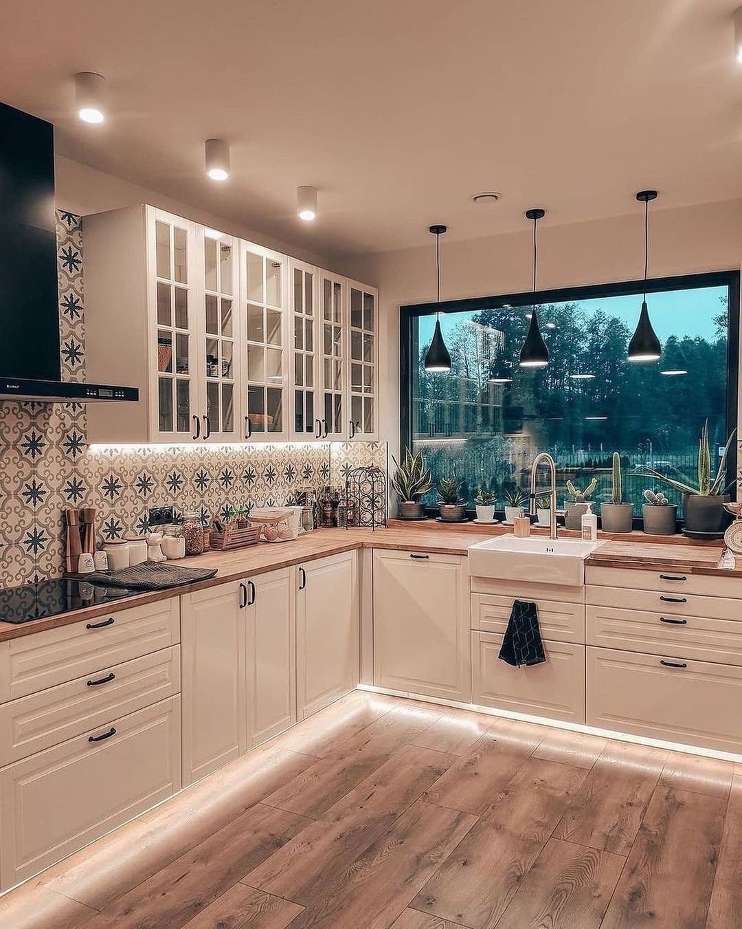 Modern farmhouse kitchen at dusk with white cabinets, wood counters, and patterned backsplash. A large, frameless picture window is filled with the dark view outside and potted succulents on the sill.
