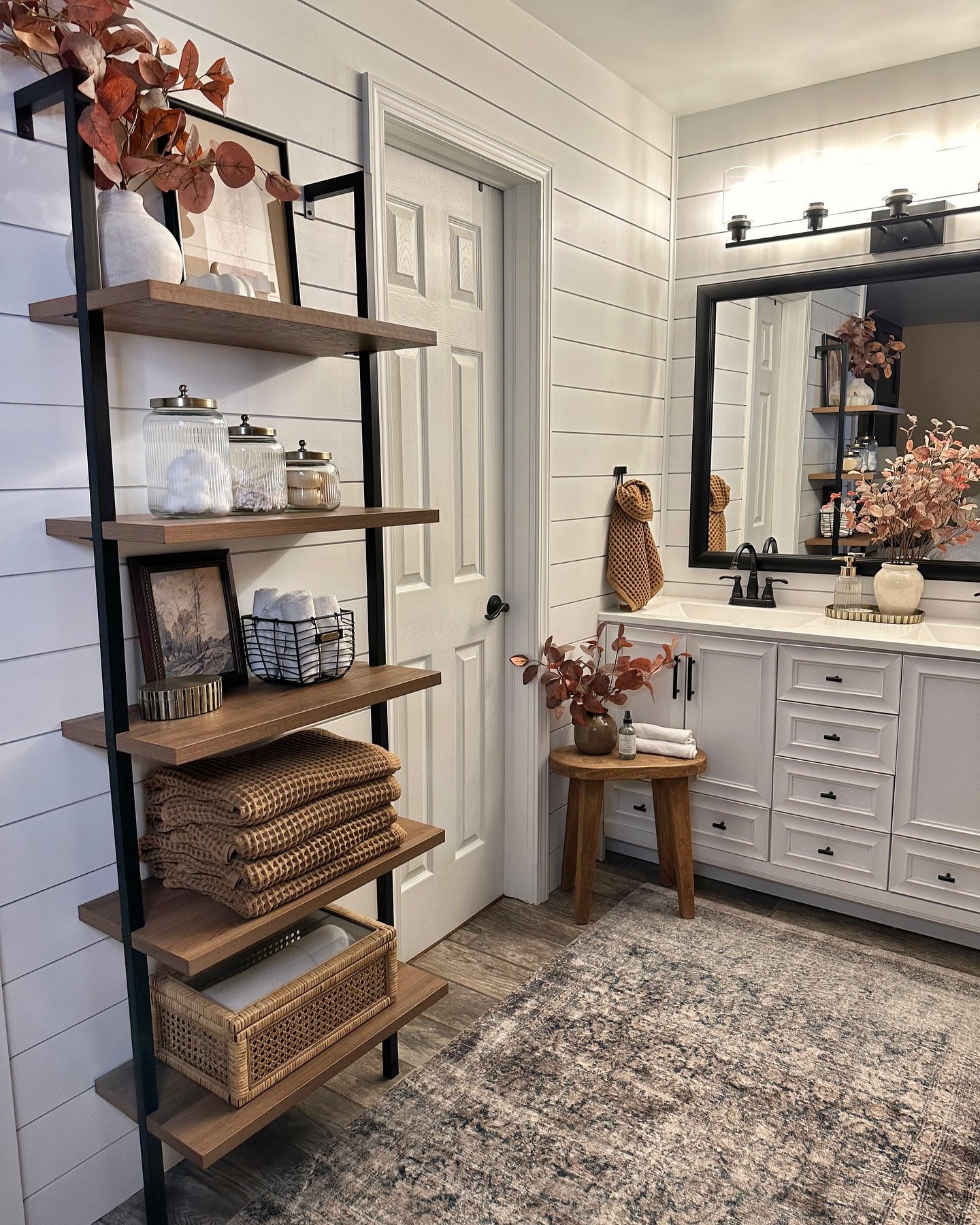 A tall, five-tier shelving unit with a black metal frame and light wood shelves, styled with folded rust-colored blankets and clear glass jars.