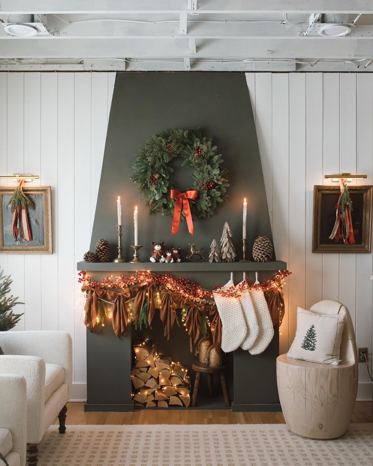 Fireplace with a dark olive green surround, decorated with a red berry garland, brown fabric swags, white knit stockings, and an evergreen wreath with a red bow.