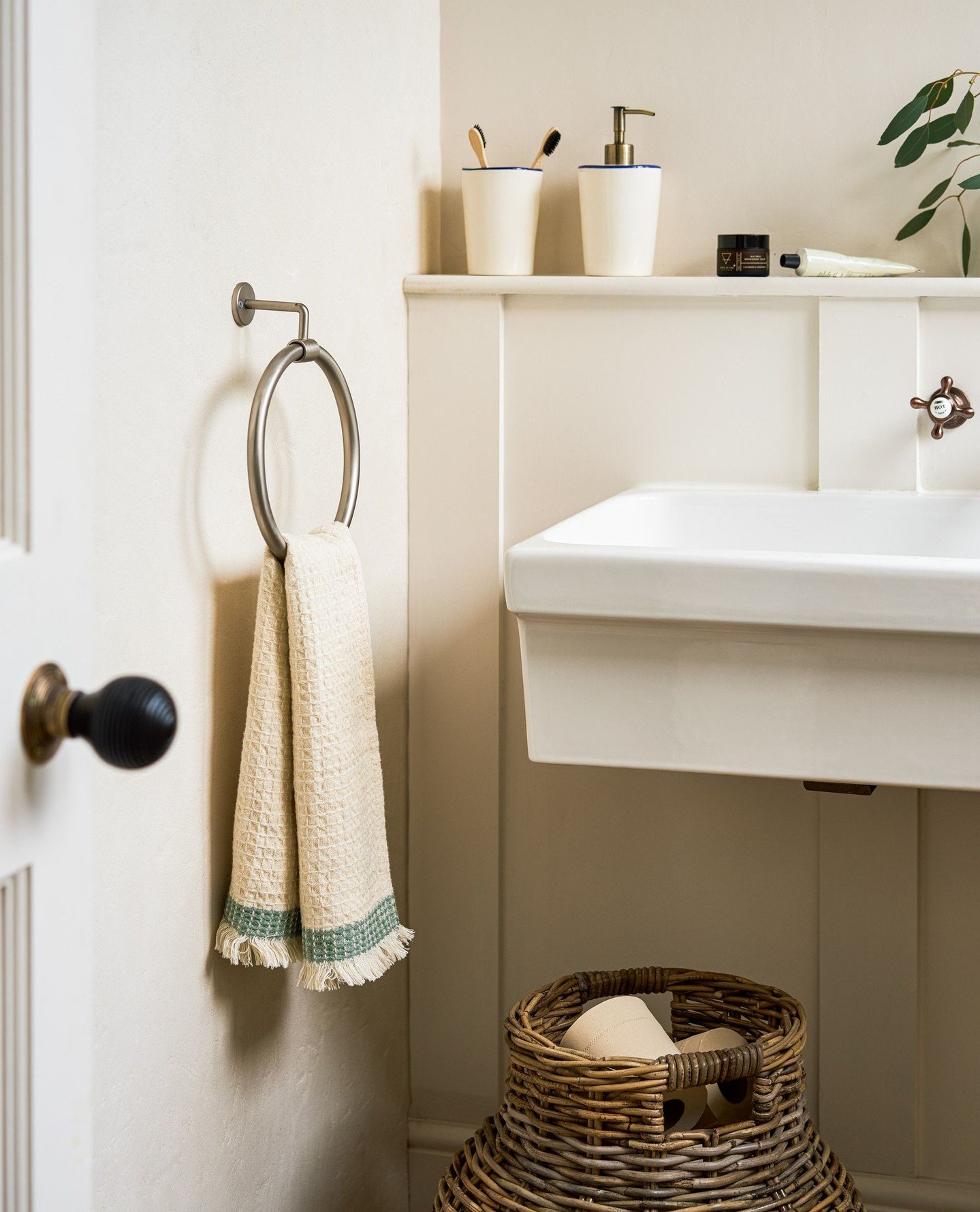 A brushed nickel metal towel ring holding a cream, fringed hand towel with a green stripe, mounted on a cream wainscoting wall.