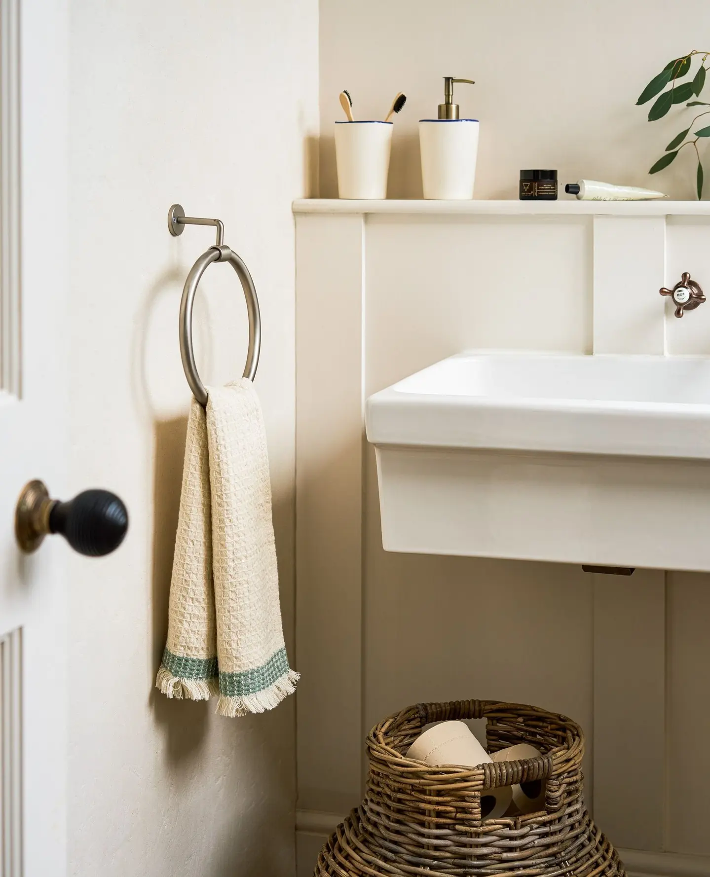 A brushed nickel metal towel ring holding a cream, fringed hand towel with a green stripe, mounted on a cream wainscoting wall.