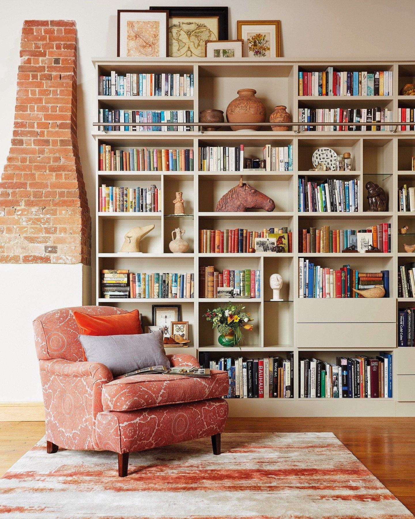 A large, custom built-in bookshelf painted a warm taupe color, filling the entire wall next to an exposed brick column, featuring a horizontal metal railing across the middle shelf and a mix of books and sculptural decor.