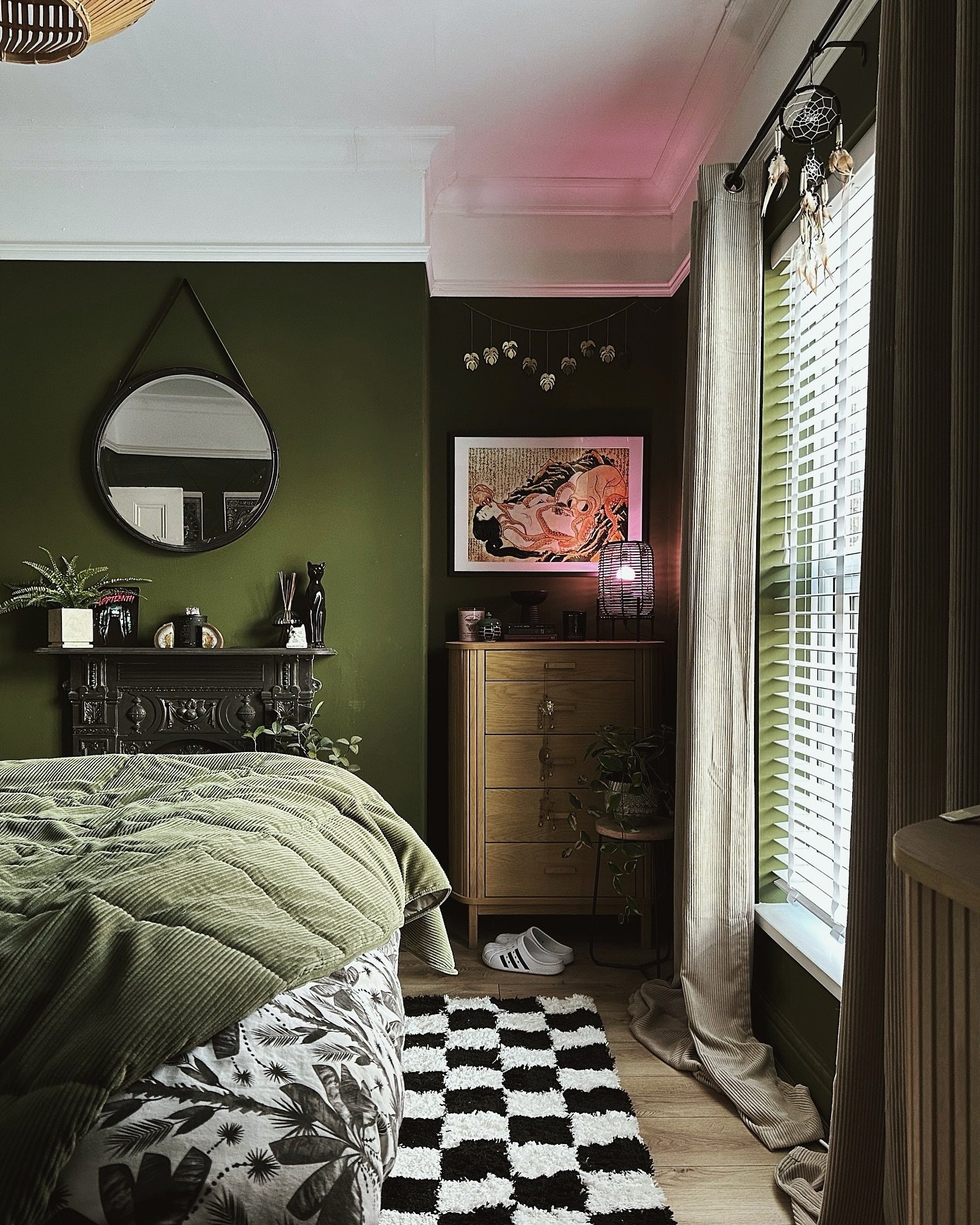 A moody, eclectic bedroom with deep olive green walls, a matching olive green comforter, a black and white checkerboard rug, and a wooden dresser.
