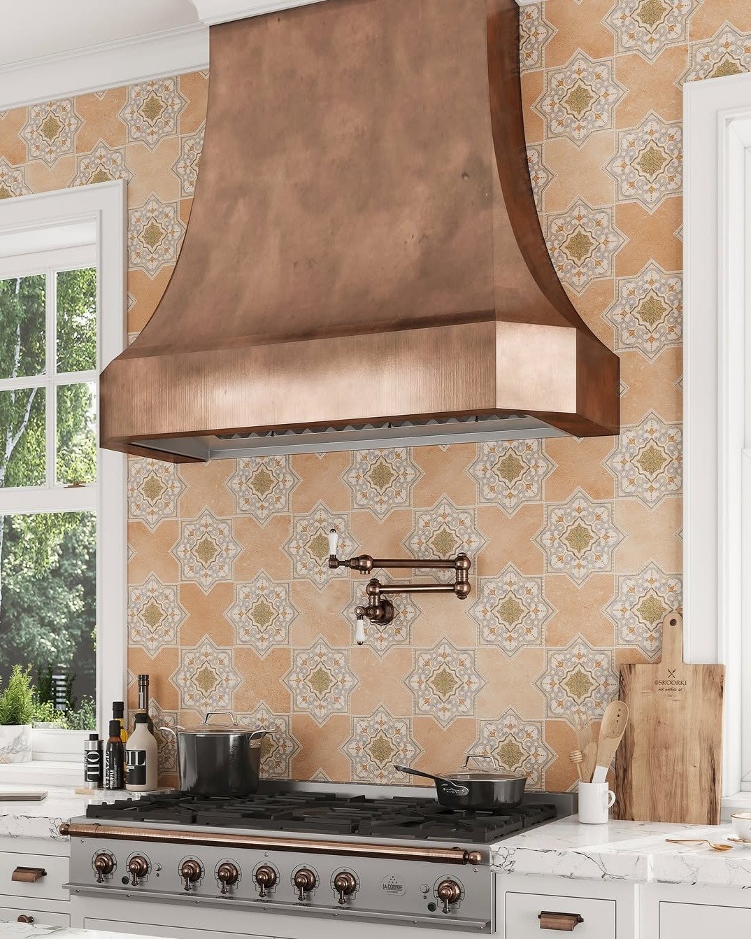 Kitchen backsplash with large square tiles featuring a terracotta, gold, and white geometric medallion pattern, set behind a hammered copper range hood.
