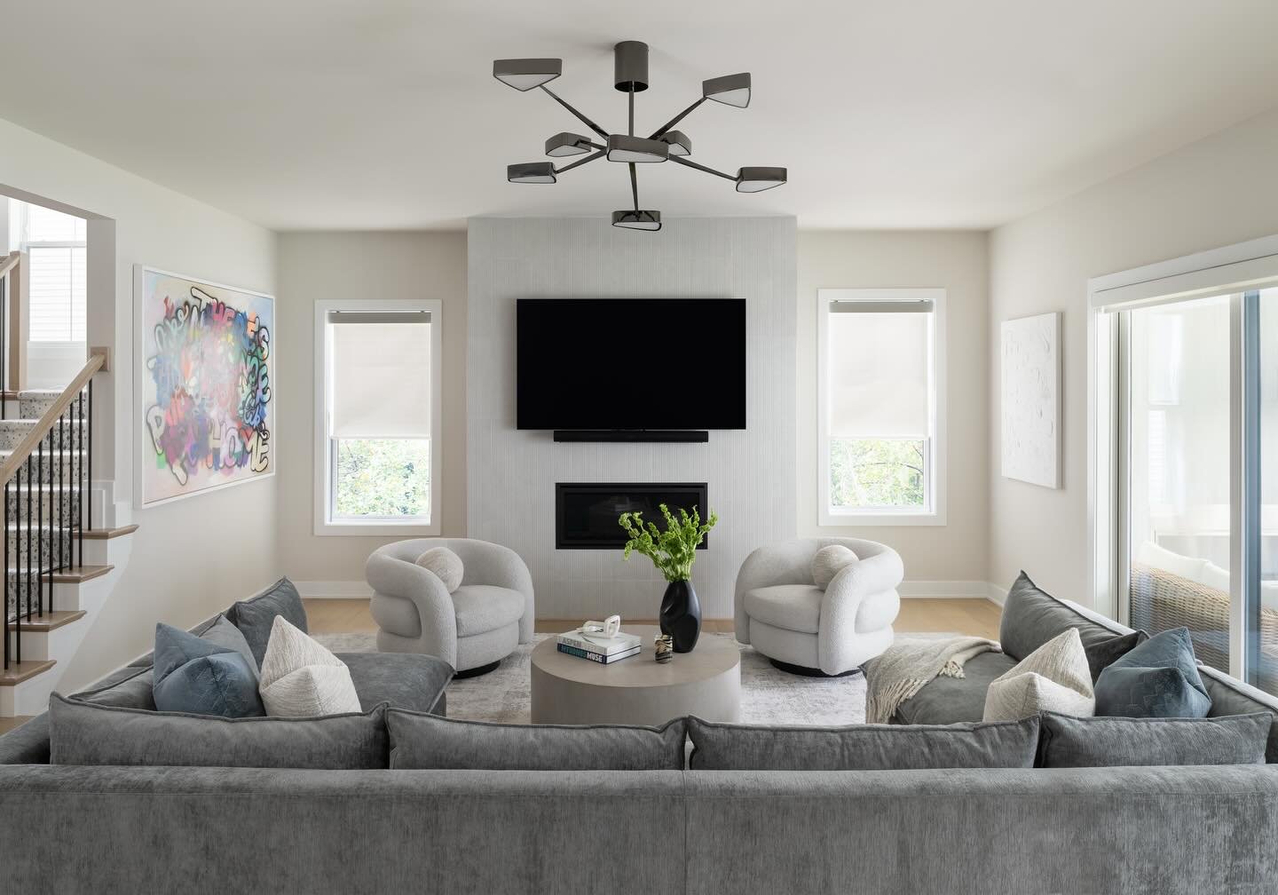 Spacious, modern living room featuring a gray sectional sofa, two white boucle swivel chairs, and a geometric chandelier above a fireplace.