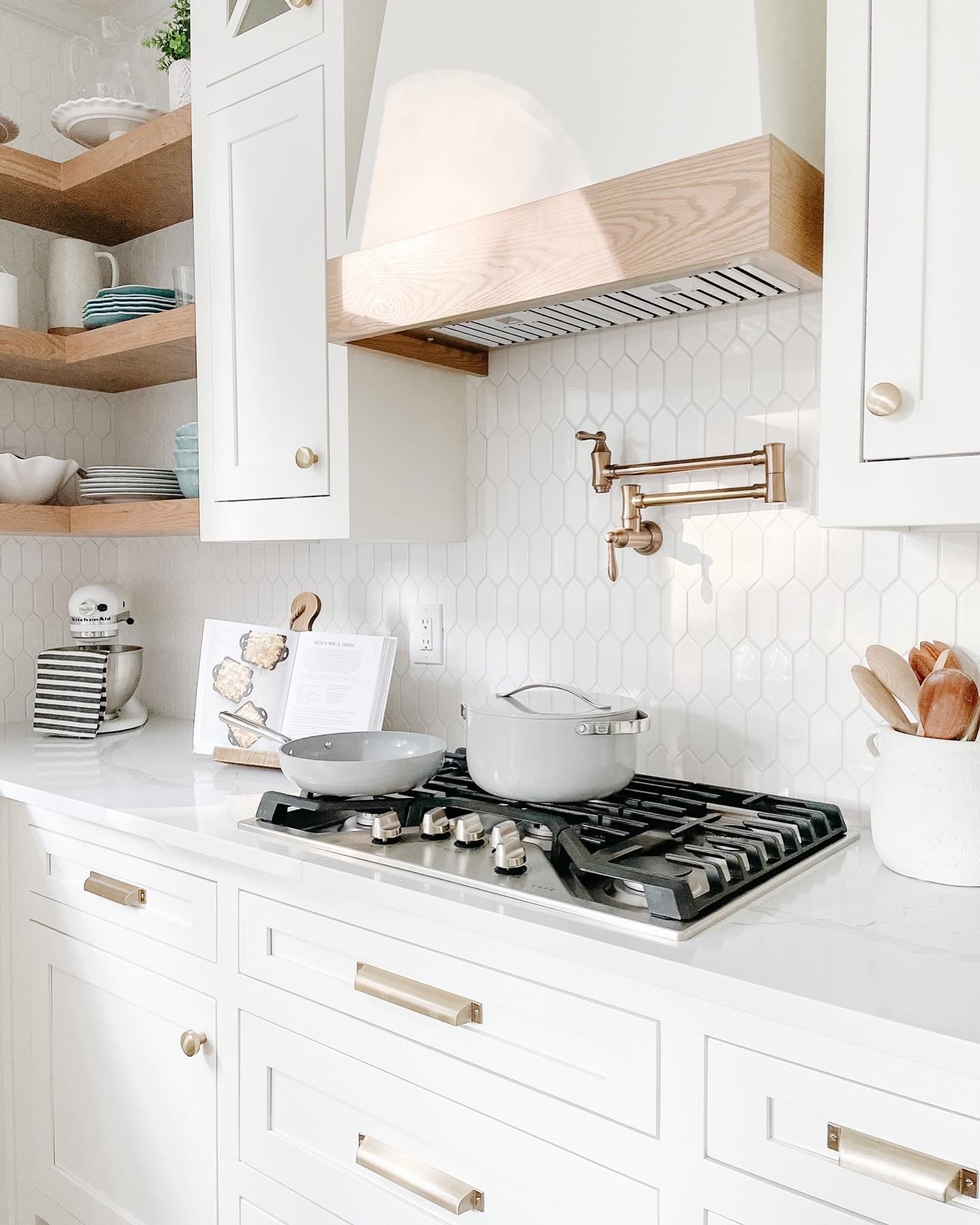 White kitchen with a white picket hexagon tile backsplash, white shaker cabinets, gold hardware, a gold pot filler, and a natural wood range hood.