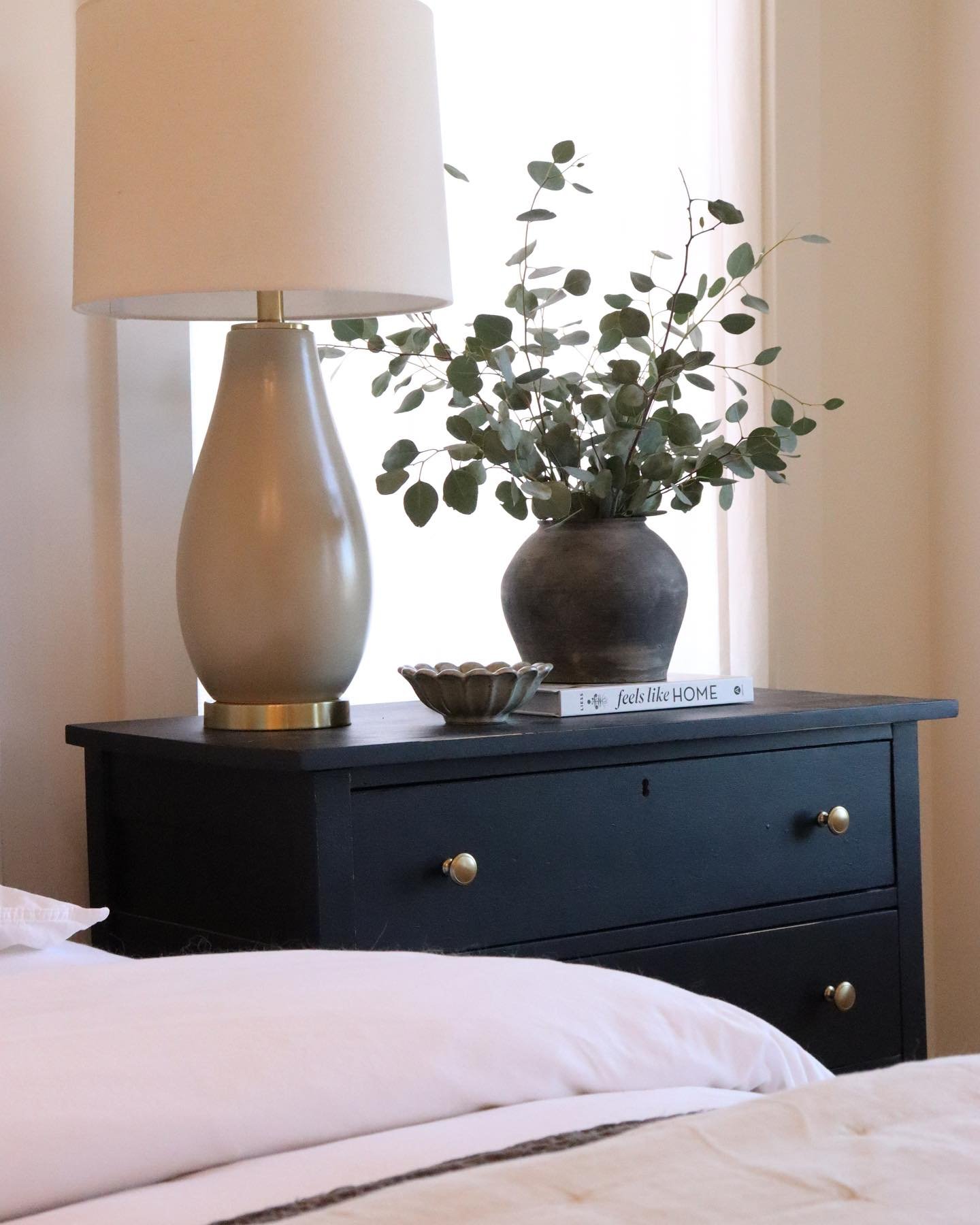 Close-up of a three-drawer black nightstand with antique brass round knobs, holding a neutral lamp and eucalyptus stems.