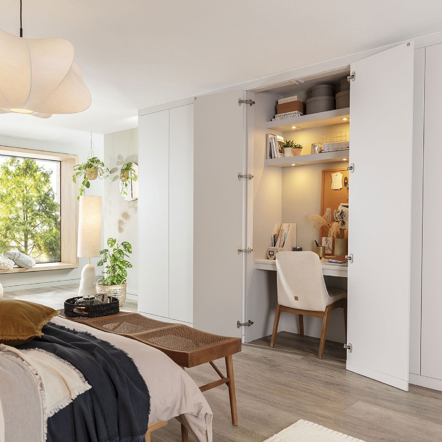 A white fitted wardrobe with two doors opened to reveal a hidden, illuminated workstation with a desk surface, shelving, and a cork board.