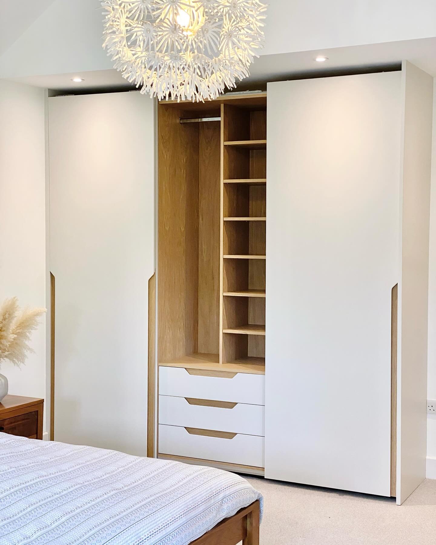 Modern wardrobe with white sliding doors, recessed oak handles, and an organized interior featuring drawers, hanging space, and adjustable wood shelves.