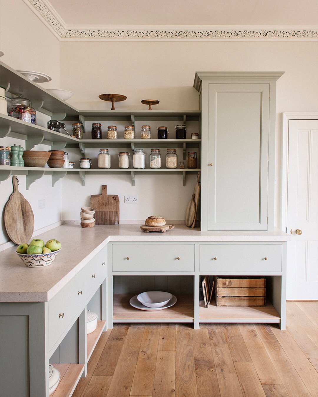 Traditional sage green kitchen with open wooden shelving holding dry goods in jars and a tall, integrated, freestanding-style storage cupboard.