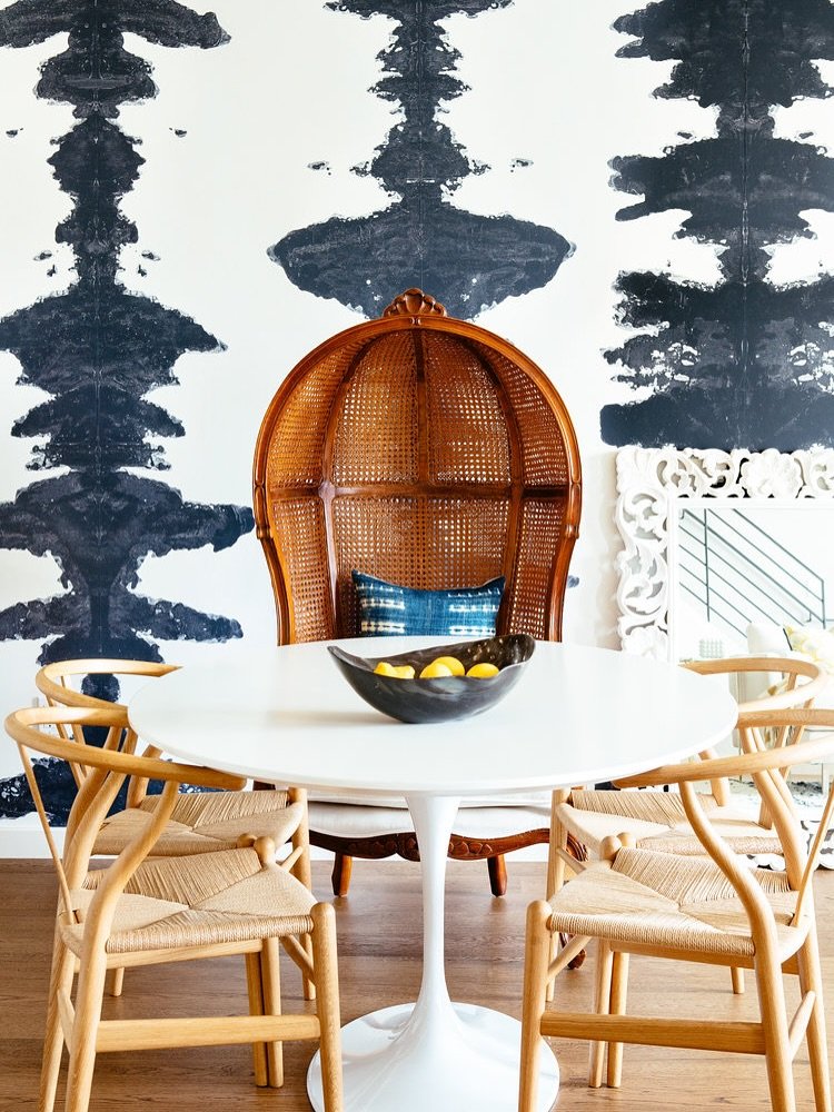A dining area with a bold black and white Rorschach-style wallpaper, a white Tulip table, natural wood Wishbone chairs, and a large, carved rattan peacock chair at the head.