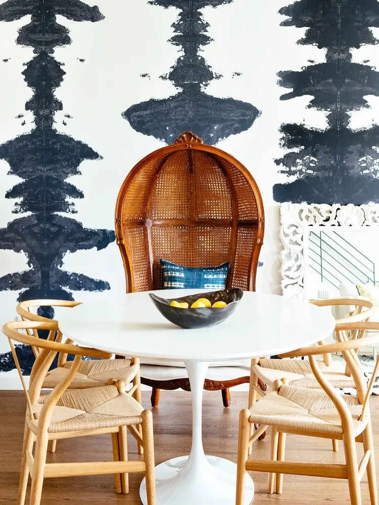 A dining area with a bold black and white Rorschach-style wallpaper, a white Tulip table, natural wood Wishbone chairs, and a large, carved rattan peacock chair at the head.