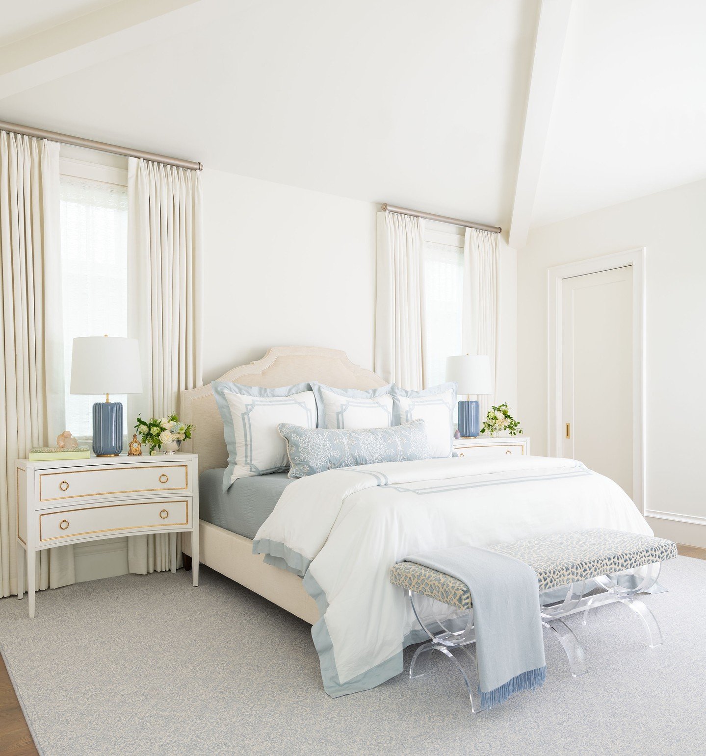 Bright, high-ceiling bedroom with a beige arched headboard, white and pale blue embroidered bedding, and two white nightstands with brass hardware and blue lamps.