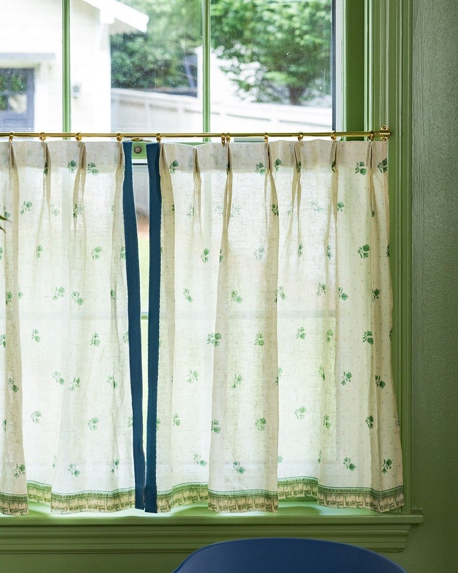 Close-up of a kitchen window with light cafe curtains featuring small green floral prints and a dark blue vertical ribbon detail, hung against a vibrant green window frame.