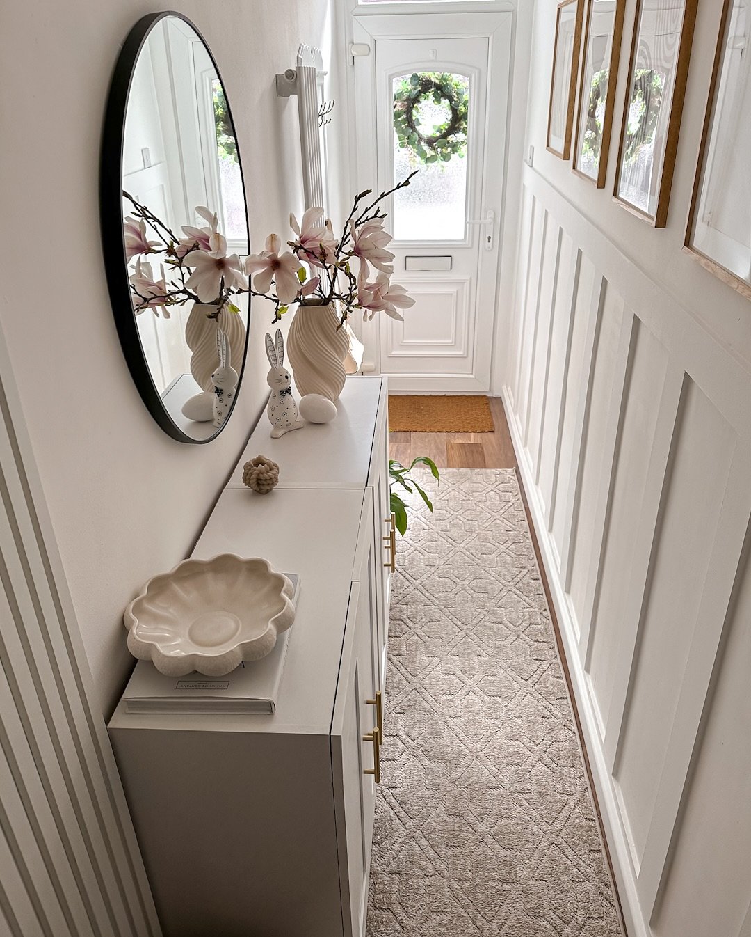 A narrow white hallway featuring tall vertical board and batten paneling, a long white storage cabinet with gold handles, a round black mirror, and a beige patterned runner
