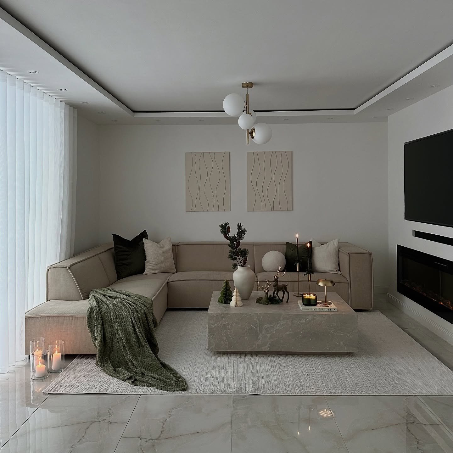 A contemporary living room featuring a beige sectional sofa, a large square marble coffee table, green velvet pillows, a knitted green throw blanket, and a thin electric fireplace.