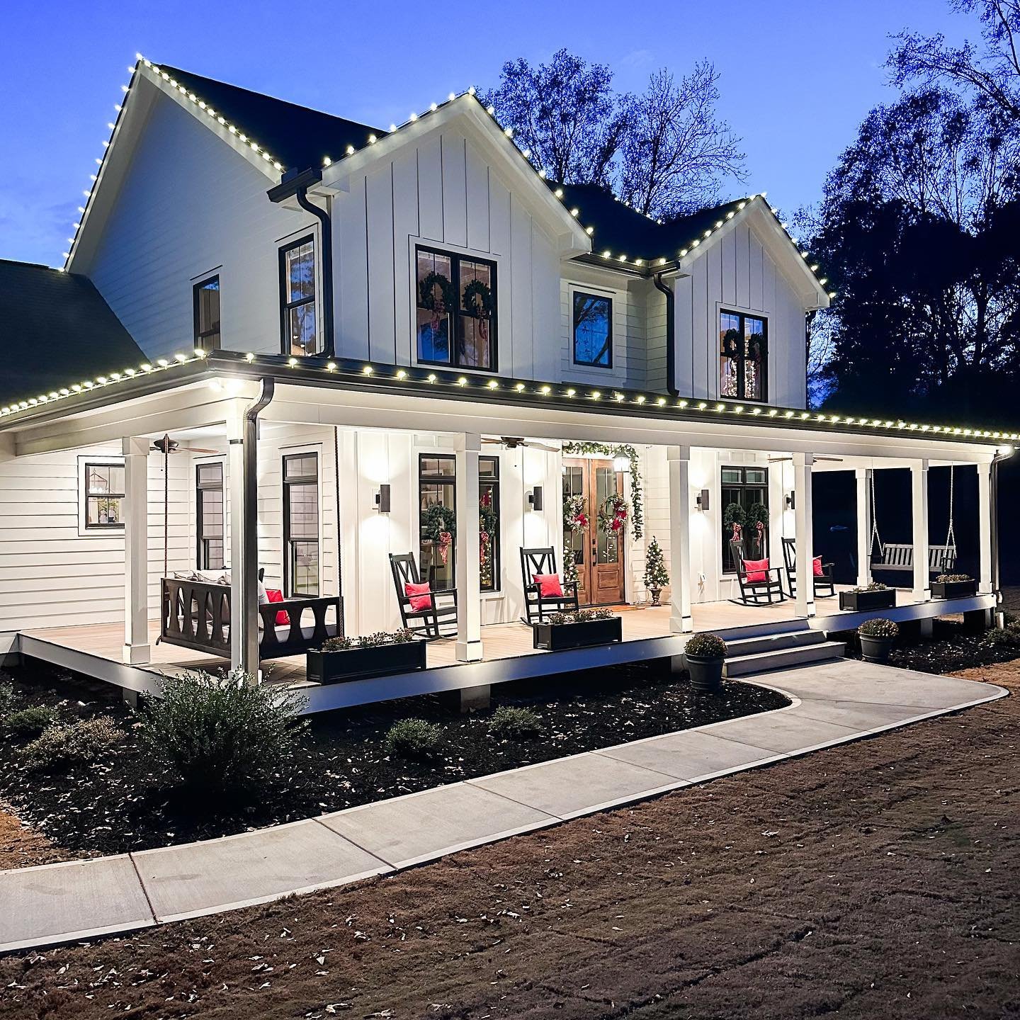 A modern white farmhouse with a large front porch is outlined with warm white LED lights along all roof edges and gables, featuring simple wreaths in the windows.