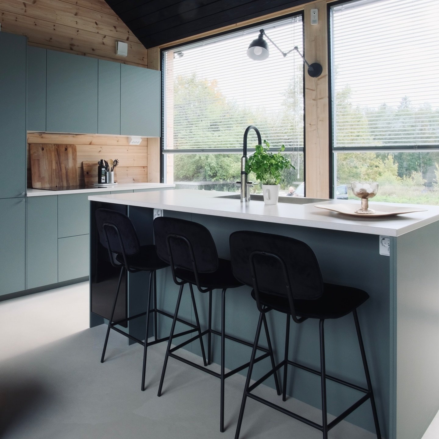 Kitchen featuring flat-front teal-colored cabinets, a white countertop island, black velvet stools, and a high, pitched ceiling lined with natural wood paneling.