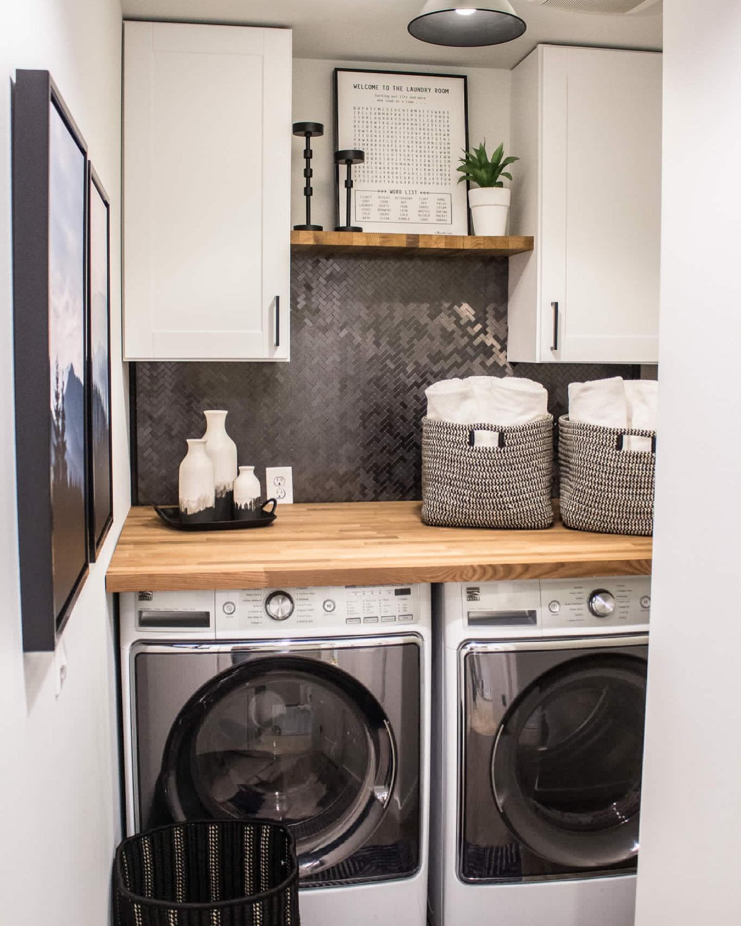 A small, modern laundry room nook featuring white upper cabinets, a light butcher block countertop over side-by-side machines, and a dramatic black metallic herringbone tile backsplash.