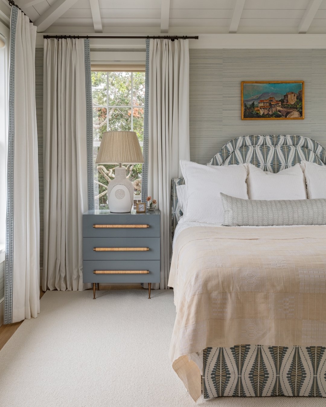 Bedroom featuring a patterned upholstered headboard and bed skirt in blue and white, a blue-gray lacquered nightstand with bamboo pulls, and fringed curtains.