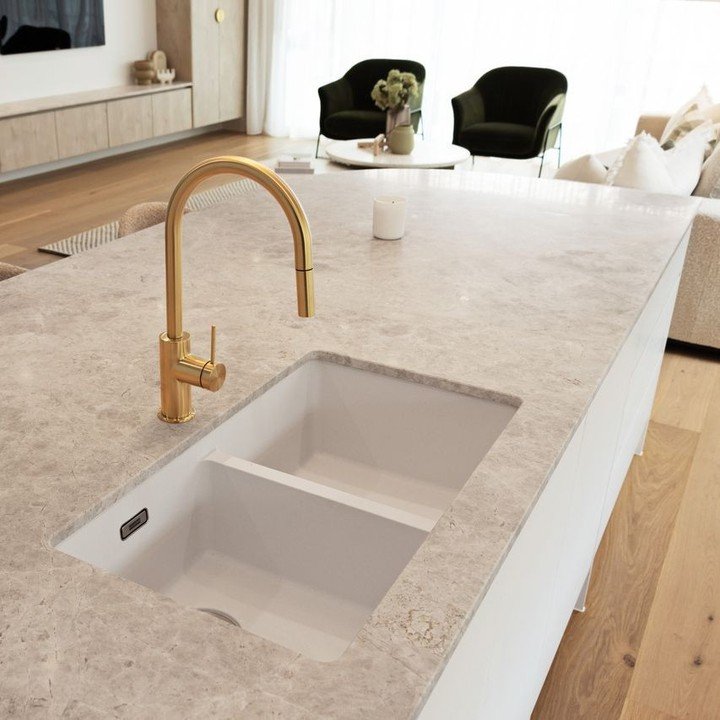 A white composite kitchen sink with a low partition (low divide) between the two bowls, installed in a light gray, marbled stone kitchen island countertop, featuring a bright gold faucet.