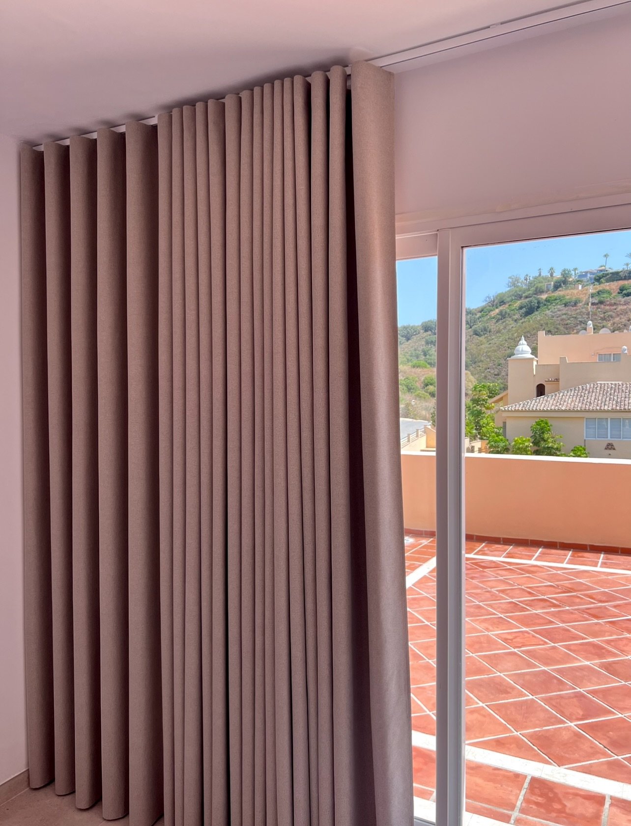 Taupe S-Fold (Wave-fold) curtains hanging from a ceiling track next to a glass patio door.