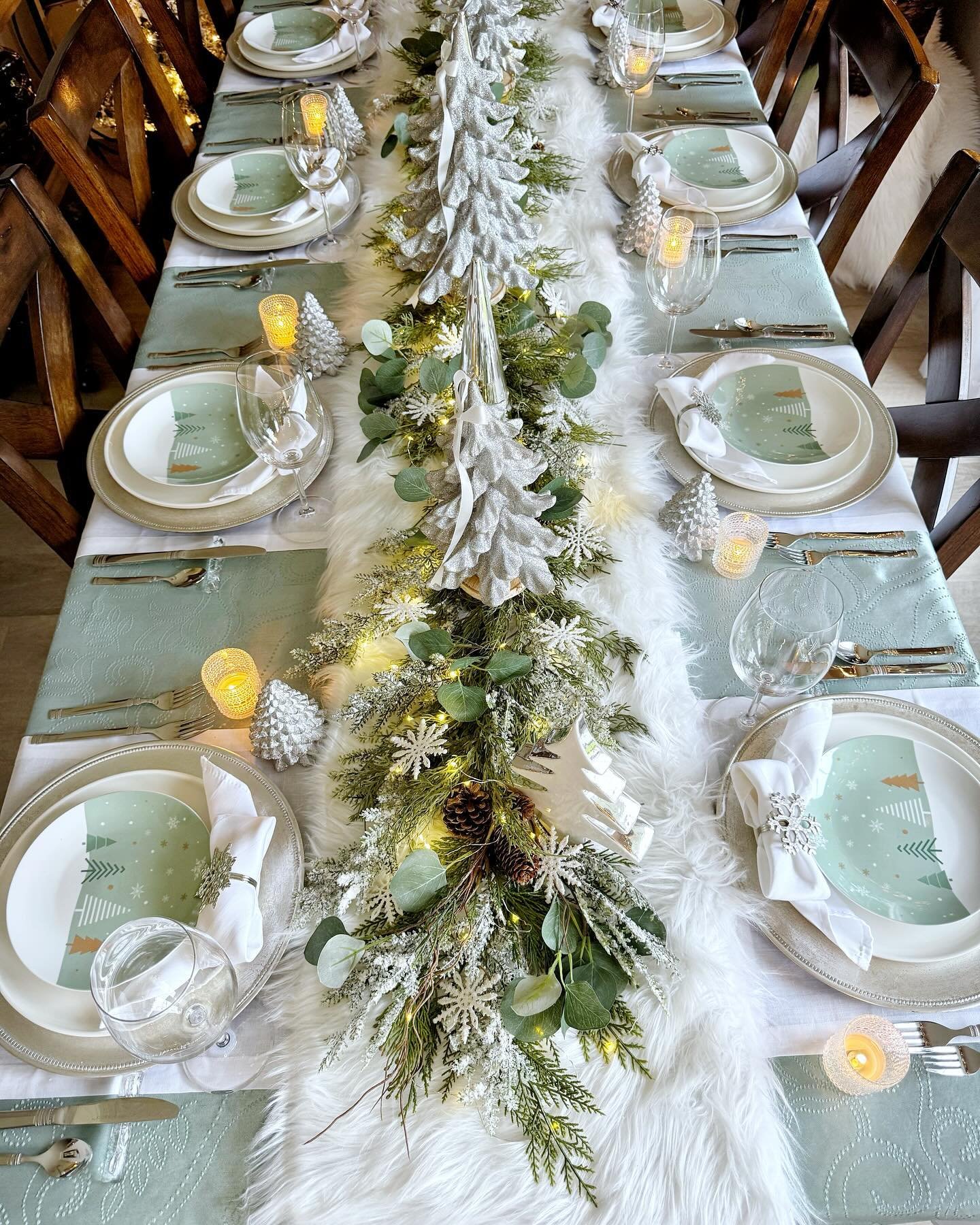 A long dining table set with light mint green placemats, silver chargers, and white plates with a modern green tree design. A centerpiece runs down the middle, featuring a white faux fur runner, snow-dusted greenery, silver glitter trees, and tiny white string lights.