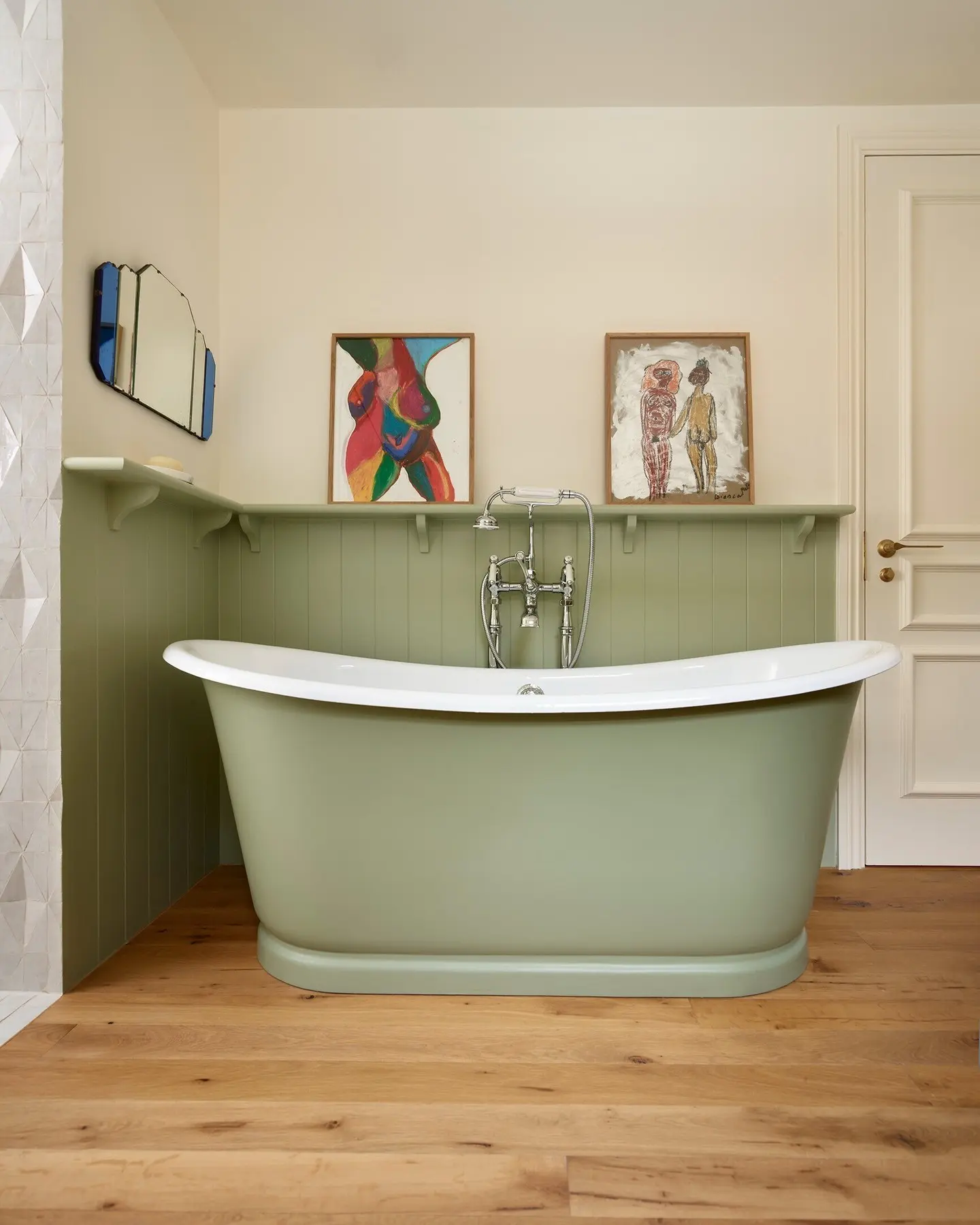 A freestanding sage-green bathtub on a light wood floor, with a green wainscoting shelf holding two framed abstract, figurative folk art paintings.