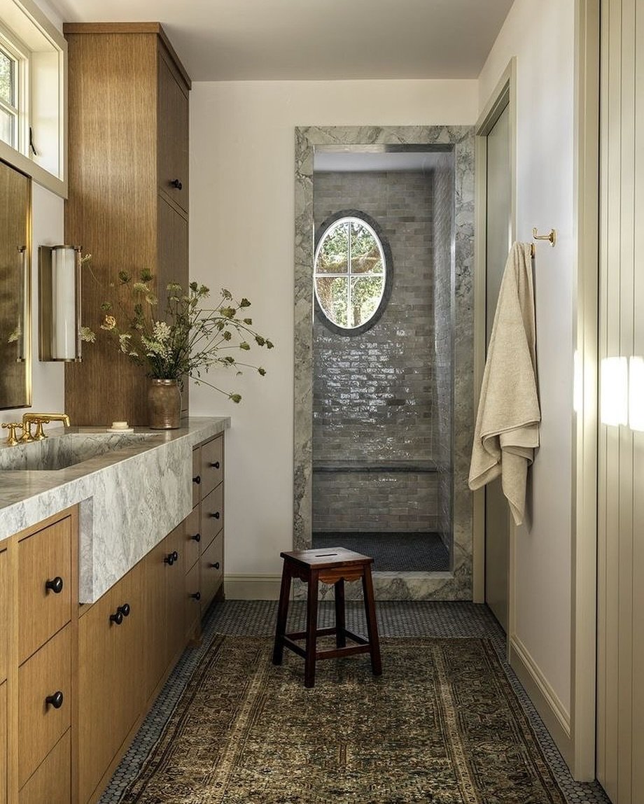 A luxury bathroom featuring light wood cabinetry, a white marble countertop, a glass shower with marble surround and an oval window, and a rectangular runner rug with a muted, earthy brown and green traditional pattern.