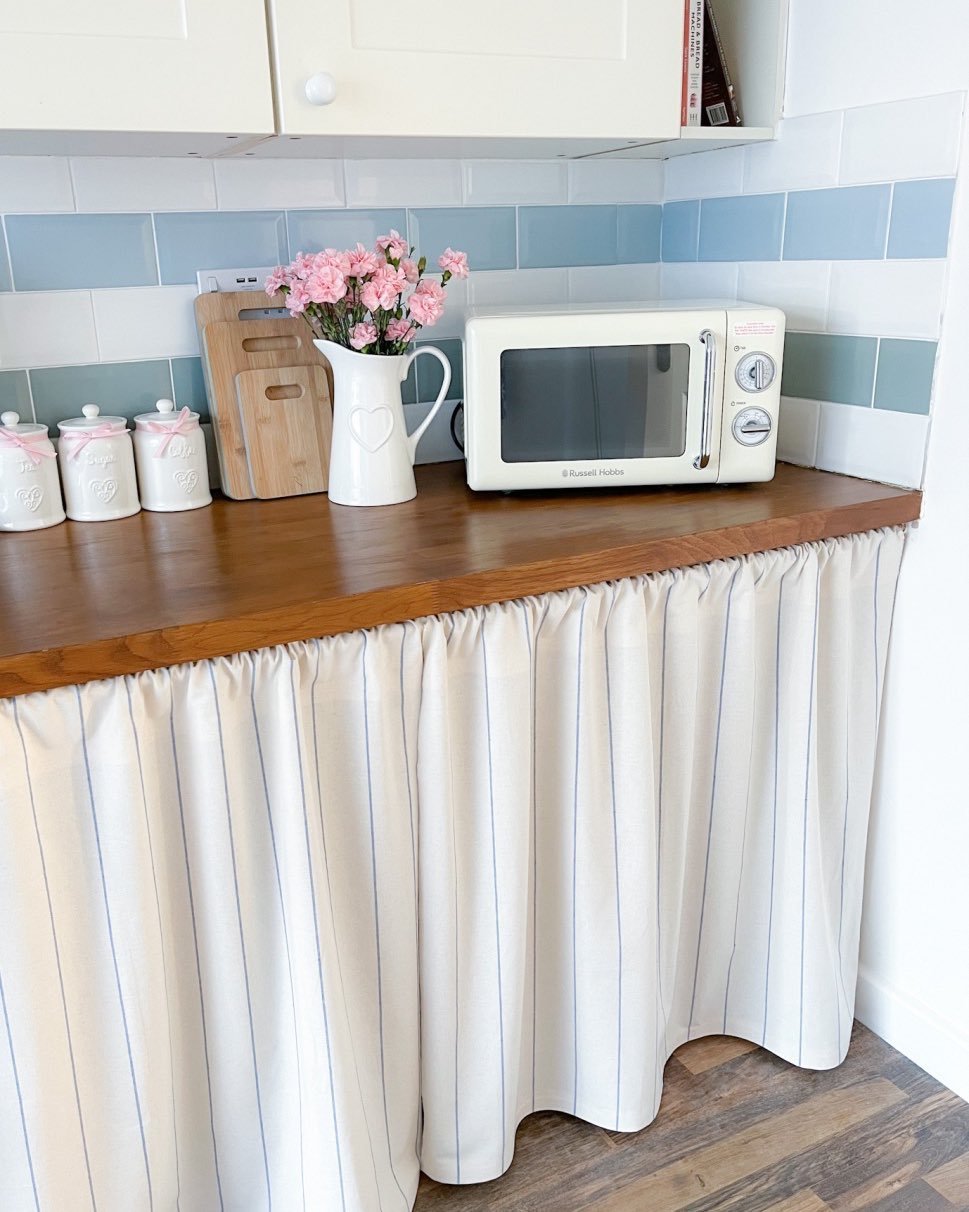 Kitchen counter with a microwave and a bouquet of pink flowers, featuring a floor-length fabric under-counter skirt in white with light blue pinstripes.