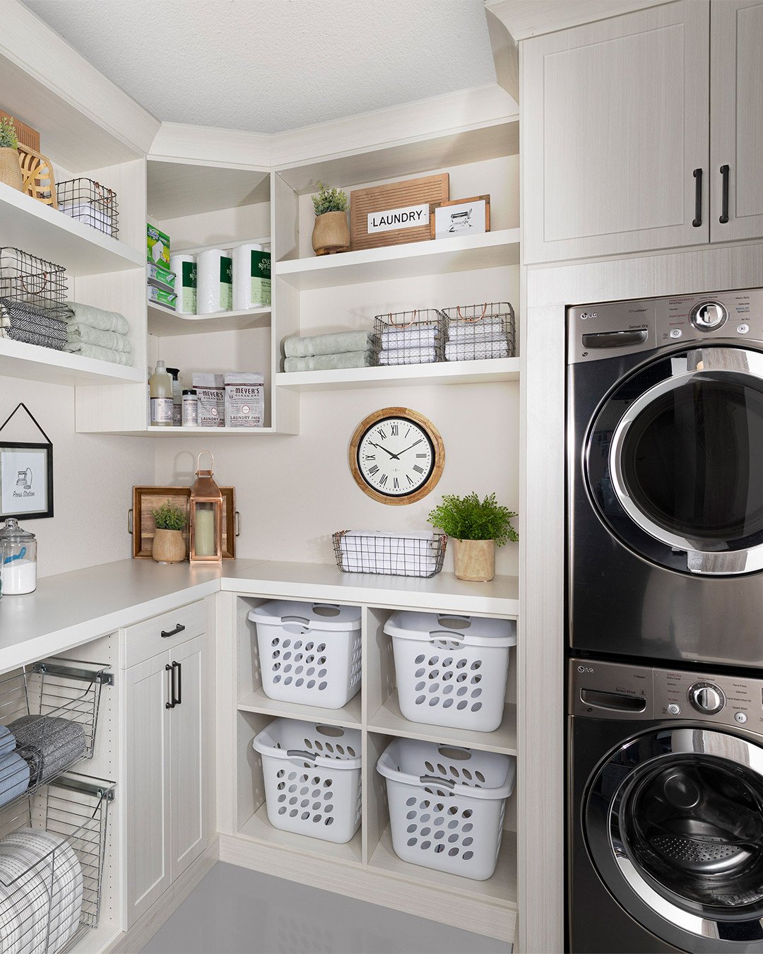 A small, highly organized white laundry room with a stacked washer and dryer unit next to a wall of custom white built-in shelving, including dedicated open cubbies for laundry hampers.