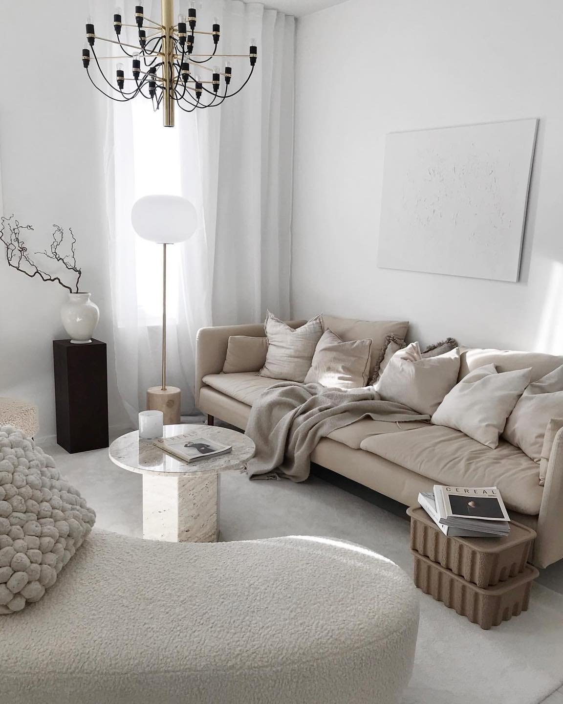 A bright, minimalist beige living room with a camel-colored sofa, a white textured painting, a black chandelier with gold accents, and a marble pedestal coffee table.
