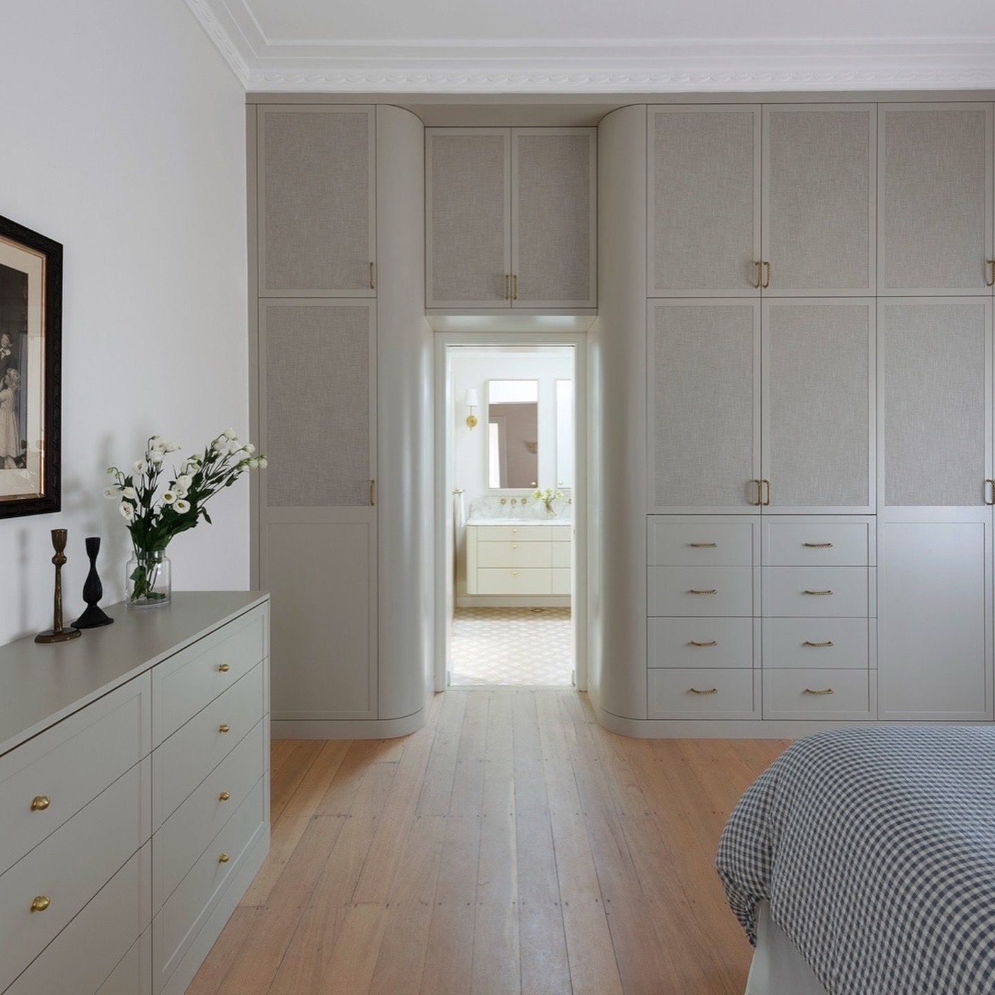 Custom light gray built-in wardrobe with textured linen door panels, brass knobs, and curved edges that frame an arched entrance to a bathroom.
