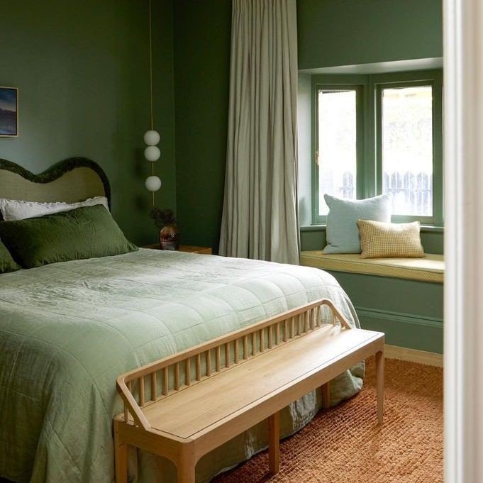 A bedroom with dark olive green walls, a lighter green quilted bedspread, a scalloped headboard, and a green window seat.