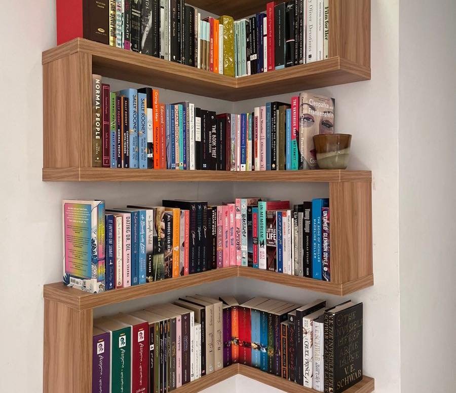 Four L-shaped, staggered floating wood shelves installed in a room corner, maximizing vertical space and filled with diverse paperbacks and hardcovers.