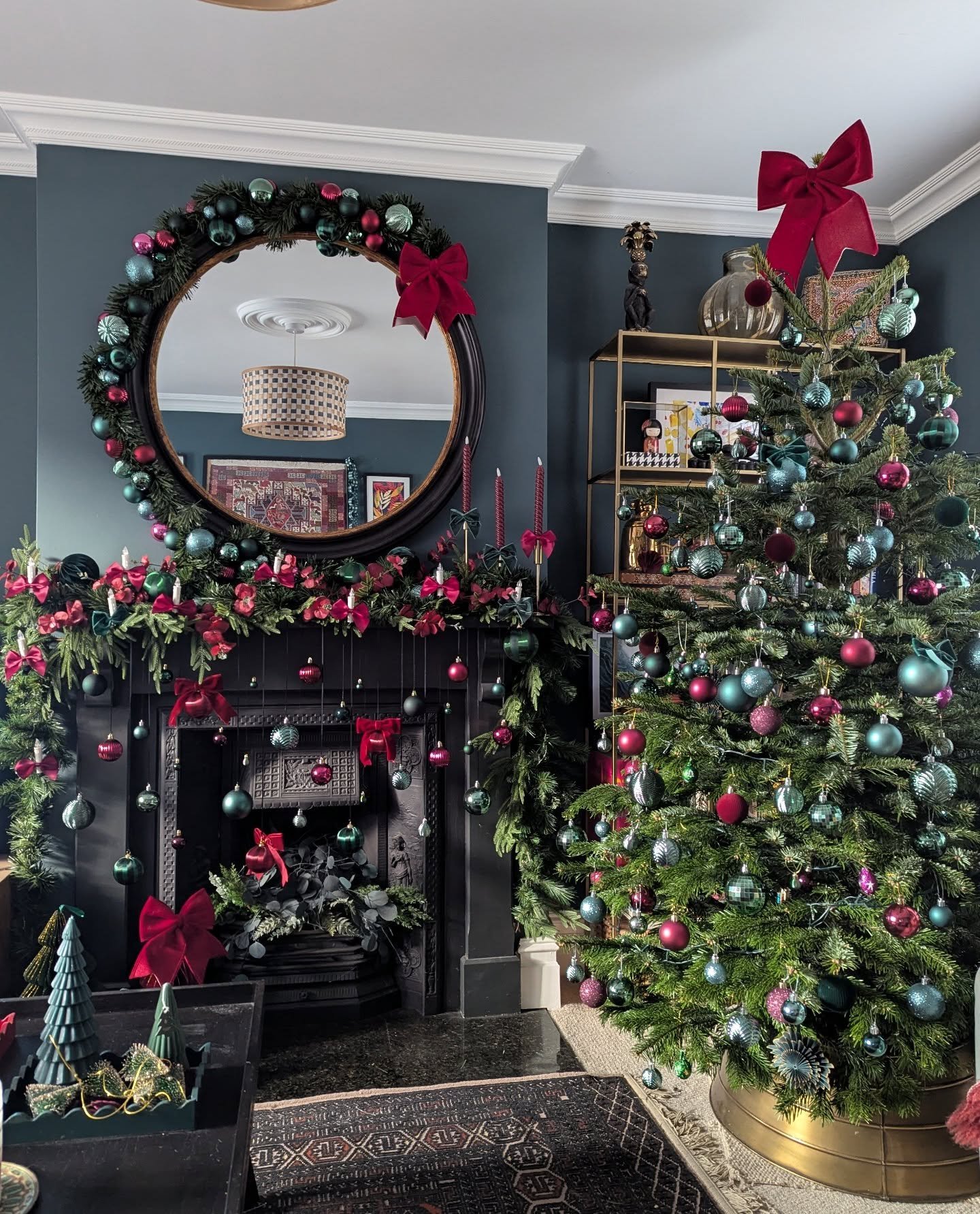 Black fireplace and dark teal wall decorated with a garland featuring deep green and teal ornaments, red ribbons, and a large round mirror framed by a wreath.
