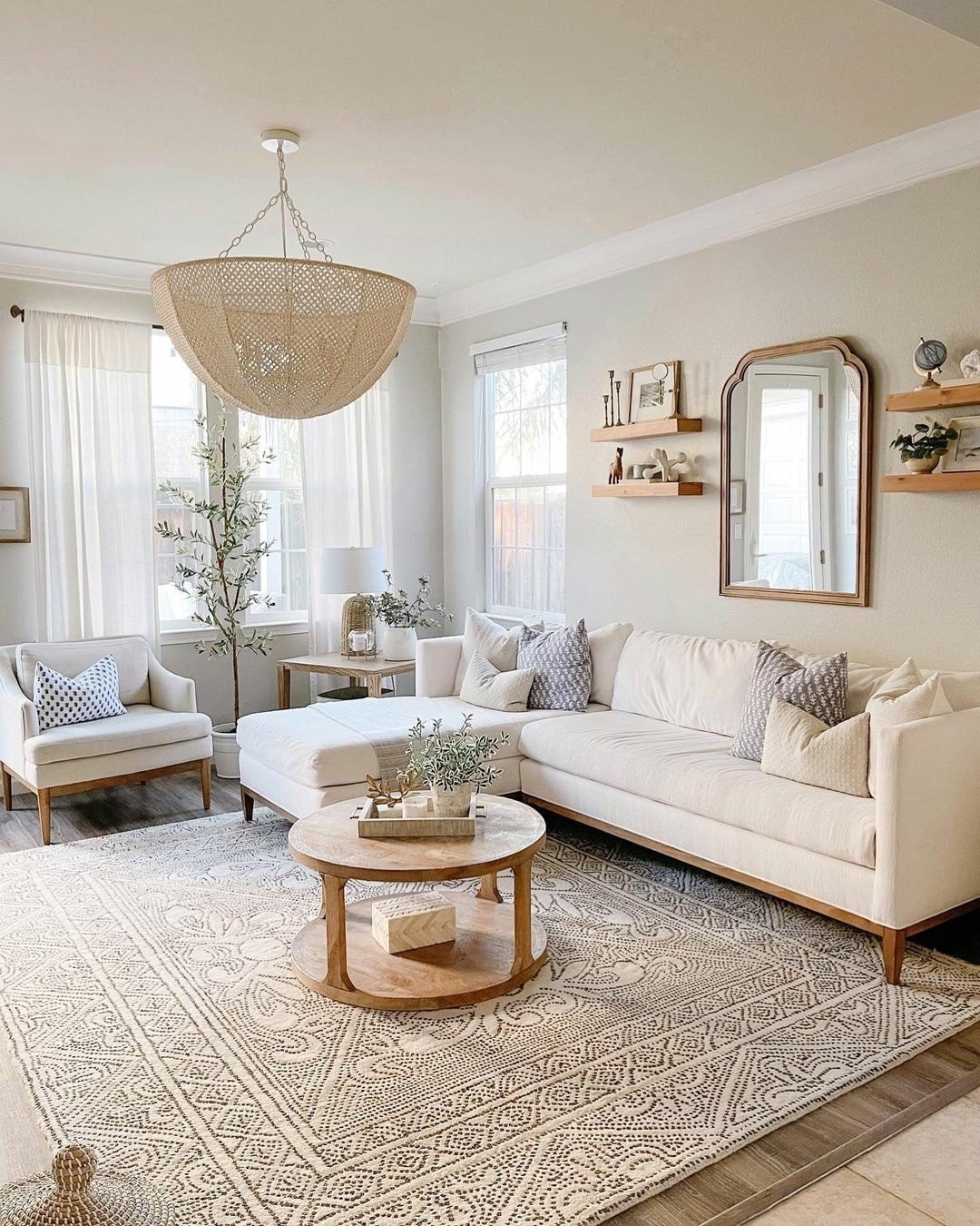 A bright neutral living room featuring a long white sofa, a light wood round coffee table, and a large woven basket-style chandelier hanging above.
